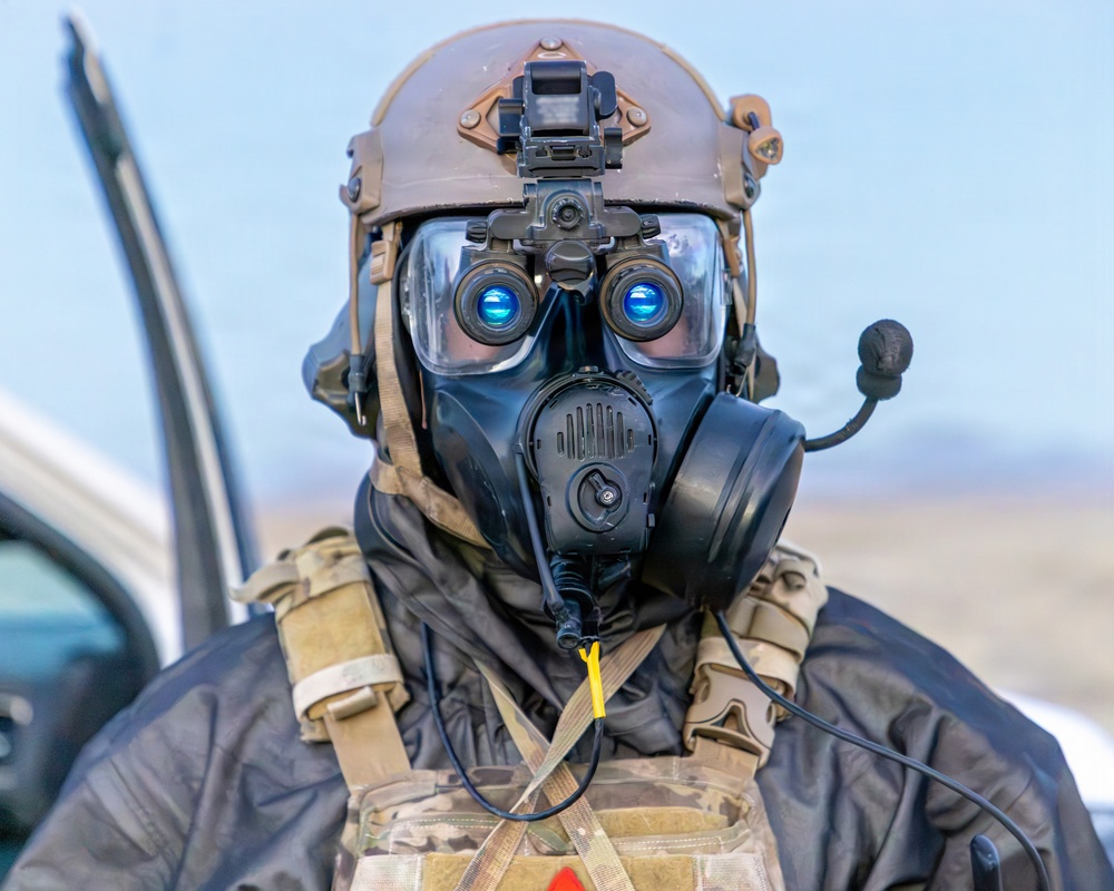 A military or tactical operator is shown wearing full CBRN (Chemical, Biological, Radiological, Nuclear) protective gear combined with advanced optical and communication equipment. The individual is equipped with a gas mask, night vision goggles mounted on a tactical helmet, and a communications microphone. The background suggests an outdoor operational or training environment near a vehicle.