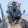 56th CRD Dugway VALEX 2026 A military or tactical operator is shown wearing full CBRN (Chemical, Biological, Radiological, Nuclear) protective gear combined with advanced optical and communication equipment. The individual is equipped with a gas mask, night vision goggles mounted on a tactical helmet, and a communications microphone. The background suggests an outdoor operational or training environment near a vehicle.
