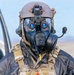 A military or tactical operator is shown wearing full CBRN (Chemical, Biological, Radiological, Nuclear) protective gear combined with advanced optical and communication equipment. The individual is equipped with a gas mask, night vision goggles mounted on a tactical helmet, and a communications microphone. The background suggests an outdoor operational or training environment near a vehicle.