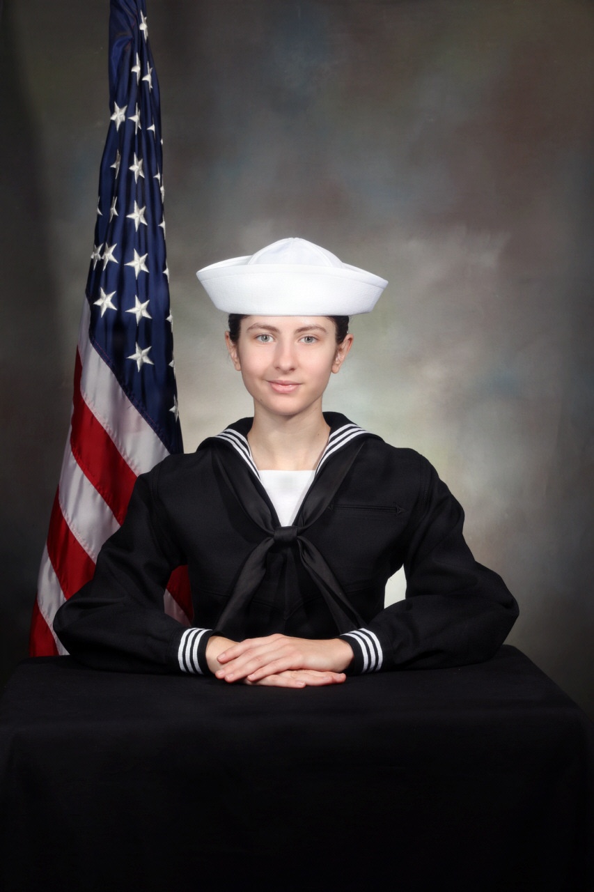 A formal military portrait of a young person wearing a U.S. Navy enlisted dress uniform (Navy Service Uniform Blue, commonly called 'Crackerjacks') with a white dixie cup cover. The subject is seated at a draped table with hands folded, posed in front of an American flag against a studio backdrop. This appears to be an official military graduation or service portrait.