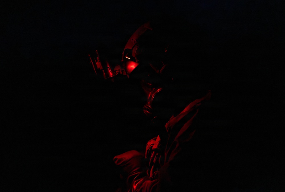 A figure wearing a gas mask and tactical gear is illuminated by a dim red light source in an otherwise completely dark environment. The low-key red lighting creates a dramatic, high-contrast silhouette effect. The subject appears to be holding or examining a small device or tool.