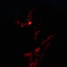 A figure wearing a gas mask and tactical gear is illuminated by a dim red light source in an otherwise completely dark environment. The low-key red lighting creates a dramatic, high-contrast silhouette effect. The subject appears to be holding or examining a small device or tool.