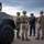 Three military personnel in OCP (Operational Camouflage Pattern) uniforms and combat gear are conversing with a law enforcement officer in a parking lot near a military vehicle. The group appears to be conducting a briefing or coordination meeting beside what is identifiable as a HMMWV (Humvee) with the marking 'WAHOTO' visible on its side. The setting appears to be an early morning or evening operation based on the overcast, dim lighting conditions.