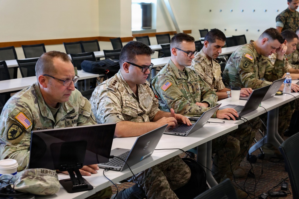 A group of military personnel in various camouflage uniforms are seated at long white tables in a classroom or training facility, working on laptops. The group includes at least one U.S. Army soldier and what appears to be soldiers from allied or partner nations based on differing uniform patterns. The setting suggests a joint military training exercise, cyber operations session, or multinational military education event.