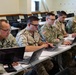 A group of military personnel in various camouflage uniforms are seated at long white tables in a classroom or training facility, working on laptops. The group includes at least one U.S. Army soldier and what appears to be soldiers from allied or partner nations based on differing uniform patterns. The setting suggests a joint military training exercise, cyber operations session, or multinational military education event.
