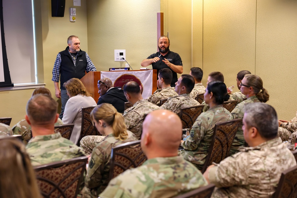 Two civilian presenters are conducting a briefing or training session for a group of military personnel in camouflage uniforms. One presenter stands at a podium while the other gestures while speaking, with an audience of approximately 15-20 soldiers seated in patterned chairs. The setting appears to be a conference or meeting room at a military installation.