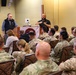 Two civilian presenters are conducting a briefing or training session for a group of military personnel in camouflage uniforms. One presenter stands at a podium while the other gestures while speaking, with an audience of approximately 15-20 soldiers seated in patterned chairs. The setting appears to be a conference or meeting room at a military installation.