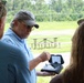 A man wearing a gray baseball cap, sunglasses, and a light blue shirt is demonstrating a handheld electronic device to two other individuals outdoors. The device appears to have a small screen displaying data or a map interface with control buttons. The setting appears to be a recreational or training area with picnic tables and a grassy field visible in the background.