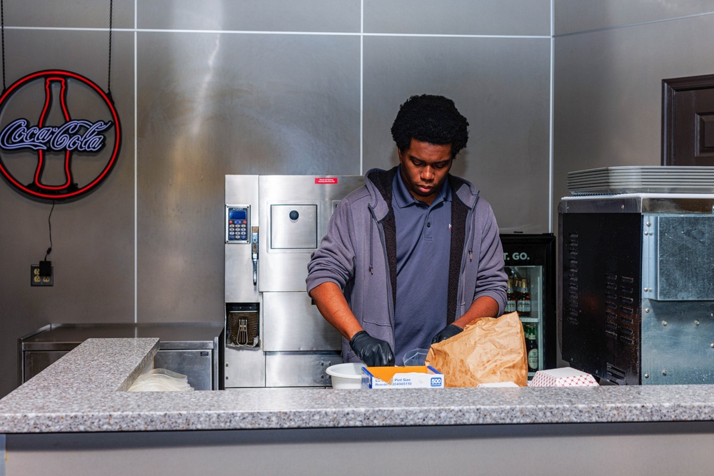 A young man wearing black gloves and a gray hoodie is working behind a food service counter, handling items including a brown paper bag and food containers. The setting appears to be a casual restaurant or food service establishment with commercial kitchen equipment visible in the background. A neon Coca-Cola sign is mounted on the wall to the left.
