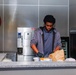 A young man wearing black gloves and a gray hoodie is working behind a food service counter, handling items including a brown paper bag and food containers. The setting appears to be a casual restaurant or food service establishment with commercial kitchen equipment visible in the background. A neon Coca-Cola sign is mounted on the wall to the left.