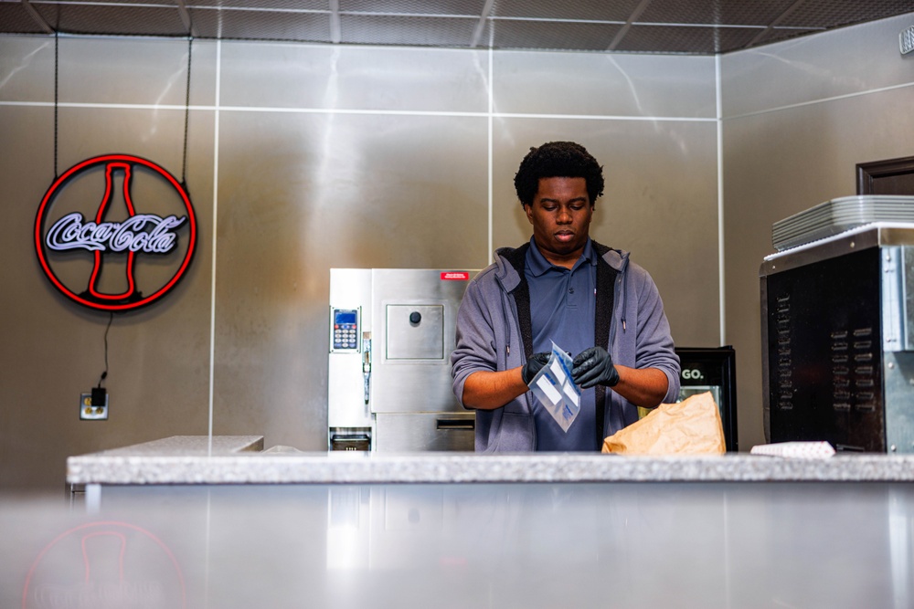 A young man wearing black gloves and a gray hoodie is working behind a service counter in what appears to be a food service or cafeteria setting. He is handling a small package or item near a brown paper bag. The environment features stainless steel surfaces, kitchen equipment, and a neon Coca-Cola sign on the wall.