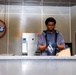 A young man wearing black gloves and a gray hoodie is working behind a service counter in what appears to be a food service or cafeteria setting. He is handling a small package or item near a brown paper bag. The environment features stainless steel surfaces, kitchen equipment, and a neon Coca-Cola sign on the wall.