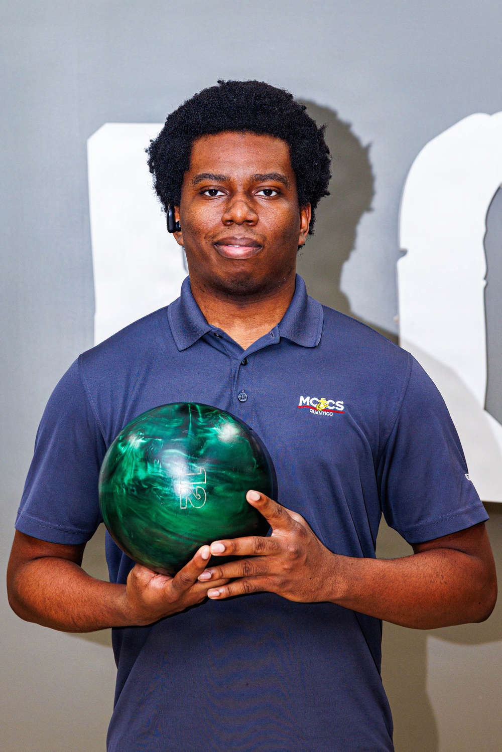 A young man poses for what appears to be a team or promotional photo while holding a green marbled bowling ball. He is wearing a navy blue polo shirt with an 'MCCS Quantico' logo on the chest. The background is a plain gray wall with partial white lettering visible.