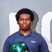 A young man poses for what appears to be a team or promotional photo while holding a green marbled bowling ball. He is wearing a navy blue polo shirt with an 'MCCS Quantico' logo on the chest. The background is a plain gray wall with partial white lettering visible.