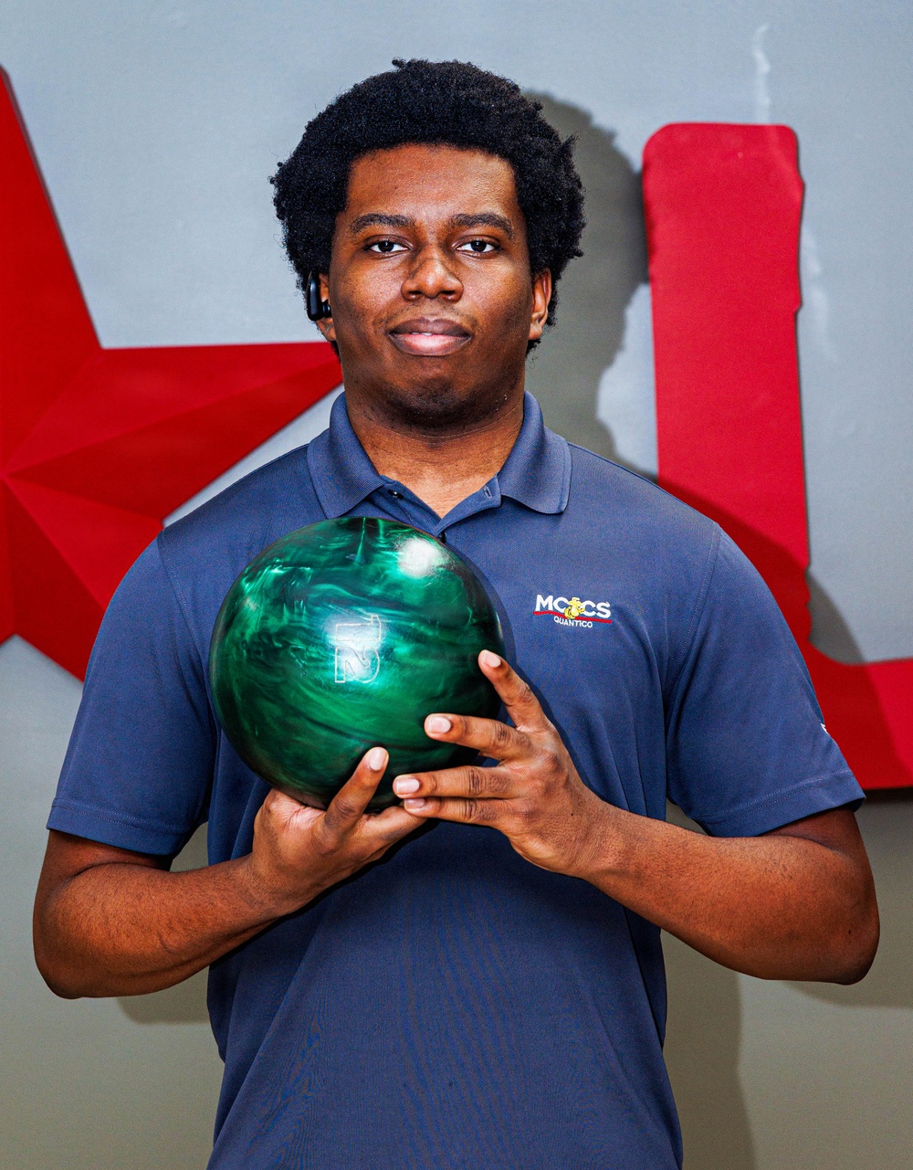 A young man is posing for what appears to be a team or promotional photo while holding a green marbled bowling ball. He is wearing a navy blue polo shirt with 'MCCS Quantico' branding. The background features a gray wall with large red decorative lettering or signage.