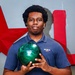 A young man is posing for what appears to be a team or promotional photo while holding a green marbled bowling ball. He is wearing a navy blue polo shirt with 'MCCS Quantico' branding. The background features a gray wall with large red decorative lettering or signage.