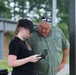 A young woman and an older man are standing outdoors, both looking down at a small electronic device the woman is holding. The man appears to be instructing or observing as the woman examines the device, which appears to be a drone controller or similar technology. They are positioned under a metal structure with trees and a building visible in the background.