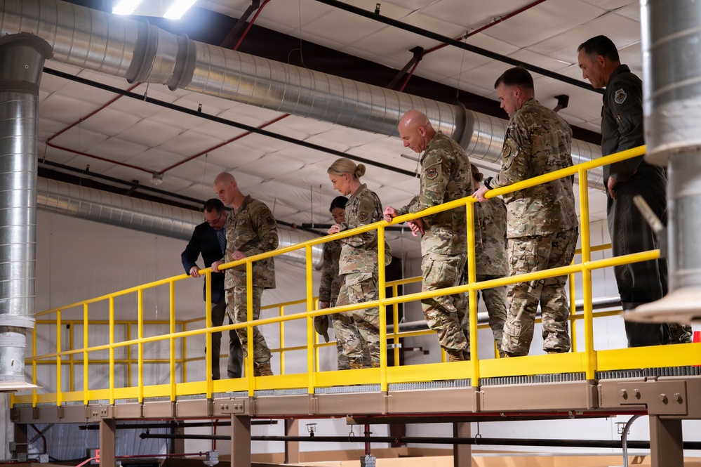 A group of military personnel in camouflage uniforms, along with one civilian in dark clothing, are walking along an elevated platform or mezzanine level inside an industrial or warehouse-type facility. The platform is bordered by bright yellow safety railings. The group appears to be conducting a facility tour or inspection.