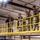A group of military personnel in camouflage uniforms, along with one civilian in dark clothing, are walking along an elevated platform or mezzanine level inside an industrial or warehouse-type facility. The platform is bordered by bright yellow safety railings. The group appears to be conducting a facility tour or inspection.