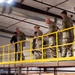 A group of military personnel in camouflage uniforms, along with one civilian in dark clothing, are walking along an elevated platform or mezzanine level inside an industrial or warehouse-type facility. The platform is bordered by bright yellow safety railings. The group appears to be conducting a facility tour or inspection.