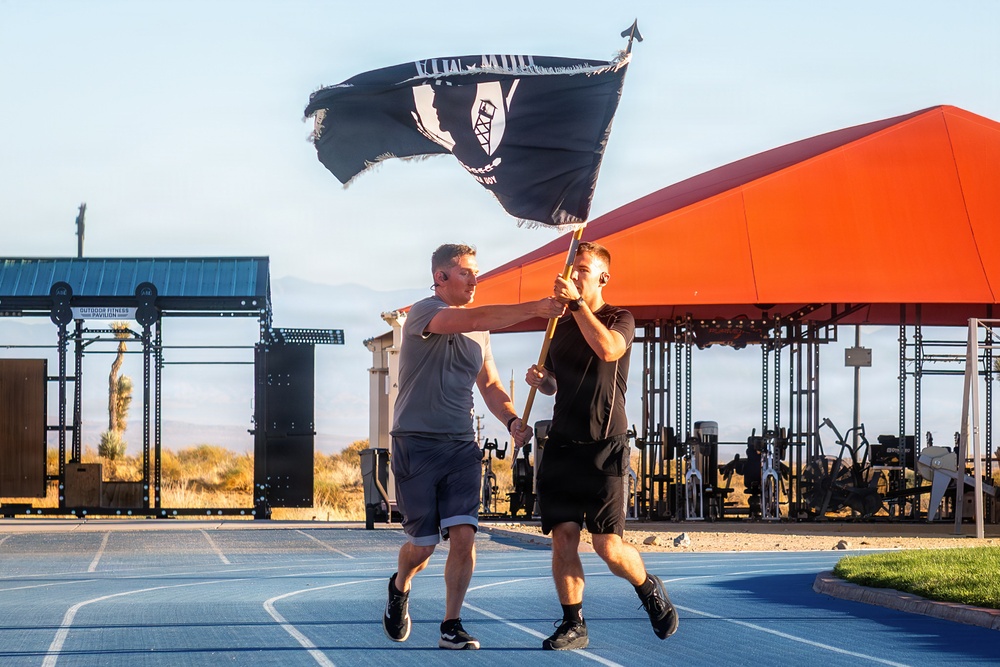Two men are carrying a large black POW/MIA flag together on a blue running track at what appears to be a military outdoor fitness facility. The scene is set during golden hour lighting, with an orange-roofed pavilion and outdoor gym equipment visible in the background. The flag is visibly worn and tattered, suggesting it has been in use for some time.