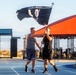 Two men are carrying a large black POW/MIA flag together on a blue running track at what appears to be a military outdoor fitness facility. The scene is set during golden hour lighting, with an orange-roofed pavilion and outdoor gym equipment visible in the background. The flag is visibly worn and tattered, suggesting it has been in use for some time.