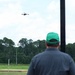 A person wearing a green cap and gray jacket is observed from behind, watching or operating a quadcopter drone flying in the sky above an open grassy field. The setting appears to be a designated drone testing or flying area, evidenced by a small landing pad visible in the lower left. Power lines and a treeline of pine trees form the background.