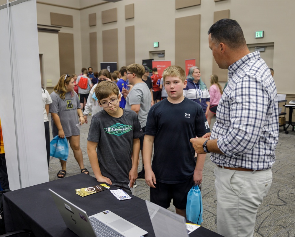 The image shows what appears to be a career fair or educational expo event held in a large convention or conference hall. Two middle-school-aged boys are interacting with an adult male presenter at a booth equipped with a laptop and printed materials. The background shows numerous other students and adults browsing additional booths throughout the venue.