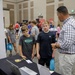 The image shows what appears to be a career fair or educational expo event held in a large convention or conference hall. Two middle-school-aged boys are interacting with an adult male presenter at a booth equipped with a laptop and printed materials. The background shows numerous other students and adults browsing additional booths throughout the venue.