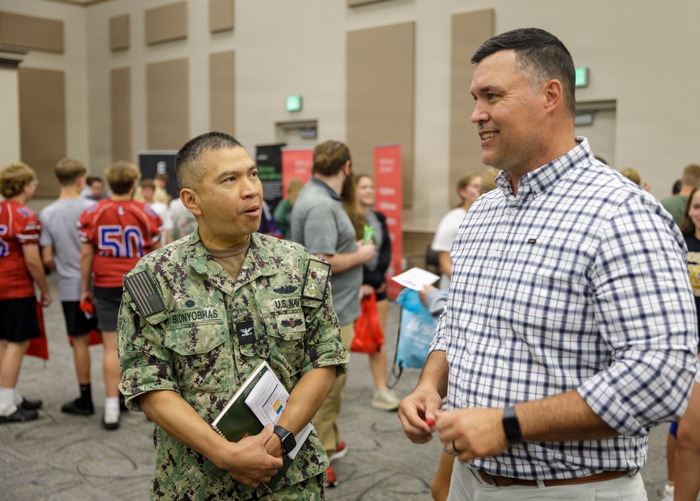 A U.S. Navy servicemember in digital camouflage uniform is engaged in conversation with a civilian man wearing a plaid shirt at what appears to be a career fair or recruitment event. The Navy personnel, identified by name tape as 'BIONYOBRAS,' holds a notebook and documents while speaking. The background shows a large indoor venue with multiple attendees, including youth in football jerseys.