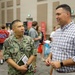 A U.S. Navy servicemember in digital camouflage uniform is engaged in conversation with a civilian man wearing a plaid shirt at what appears to be a career fair or recruitment event. The Navy personnel, identified by name tape as 'BIONYOBRAS,' holds a notebook and documents while speaking. The background shows a large indoor venue with multiple attendees, including youth in football jerseys.