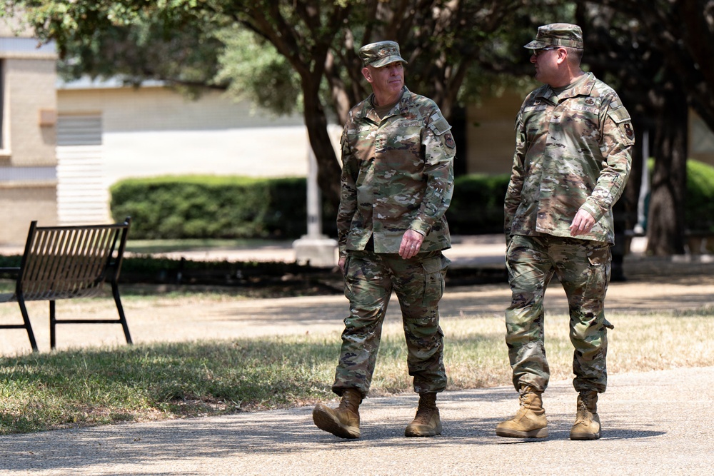 Two military personnel in U.S. Army OCP (Operational Camouflage Pattern) uniforms are walking and conversing on a paved path on what appears to be a military installation or base. Both individuals are wearing matching camouflage uniforms, patrol caps, and tan combat boots. The setting includes trees, manicured shrubs, a park bench, and institutional buildings consistent with a military campus environment.