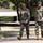 Two military personnel in U.S. Army OCP (Operational Camouflage Pattern) uniforms are walking and conversing on a paved path on what appears to be a military installation or base. Both individuals are wearing matching camouflage uniforms, patrol caps, and tan combat boots. The setting includes trees, manicured shrubs, a park bench, and institutional buildings consistent with a military campus environment.