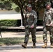 Two military personnel in U.S. Army OCP (Operational Camouflage Pattern) uniforms are walking and conversing on a paved path on what appears to be a military installation or base. Both individuals are wearing matching camouflage uniforms, patrol caps, and tan combat boots. The setting includes trees, manicured shrubs, a park bench, and institutional buildings consistent with a military campus environment.