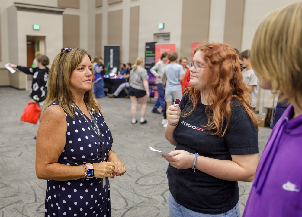 A woman wearing a navy polka-dot dress and lanyard appears to be speaking with two younger individuals at what looks like a college fair, career expo, or student recruitment event held in a large convention hall or ballroom. The foreground conversation appears to be between an adult representative and students. The background shows numerous attendees browsing various booths and displays.