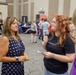 A woman wearing a navy polka-dot dress and lanyard appears to be speaking with two younger individuals at what looks like a college fair, career expo, or student recruitment event held in a large convention hall or ballroom. The foreground conversation appears to be between an adult representative and students. The background shows numerous attendees browsing various booths and displays.