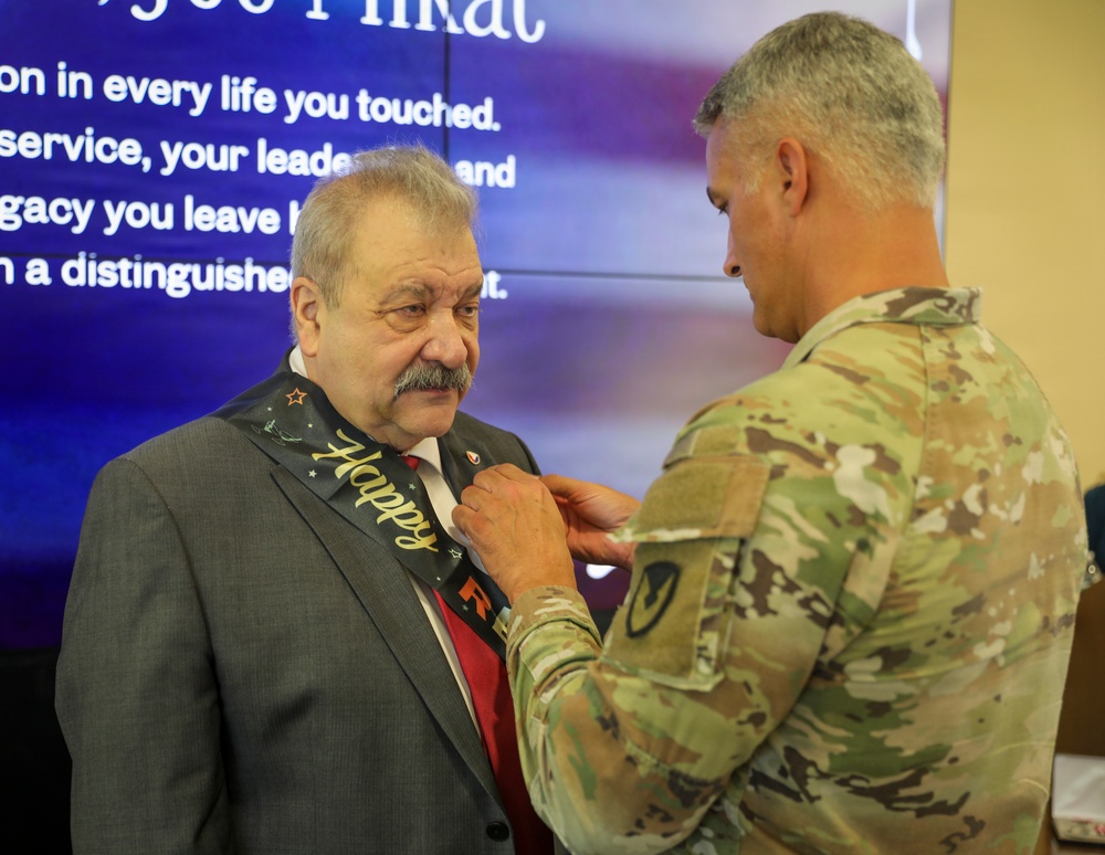 A military service member in Army Combat Uniform (ACU/OCP camouflage) is pinning or adjusting a 'Happy Retirement' sash on an older civilian man in a gray suit with a red tie. The background displays a presentation slide with text referencing distinguished service and legacy, consistent with a retirement ceremony. The event appears to be a formal military retirement celebration.