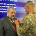 A military service member in Army Combat Uniform (ACU/OCP camouflage) is pinning or adjusting a 'Happy Retirement' sash on an older civilian man in a gray suit with a red tie. The background displays a presentation slide with text referencing distinguished service and legacy, consistent with a retirement ceremony. The event appears to be a formal military retirement celebration.
