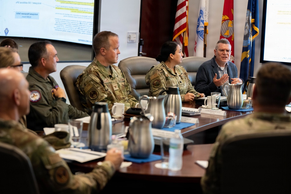 A formal military briefing or conference is taking place in what appears to be a command conference room. Several uniformed military personnel in OCP (Operational Camouflage Pattern) uniforms are seated at a large wooden conference table alongside at least one civilian. A presentation is displayed on a screen in the background, and multiple national and military branch flags are visible.