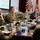 A formal military briefing or conference is taking place in what appears to be a command conference room. Several uniformed military personnel in OCP (Operational Camouflage Pattern) uniforms are seated at a large wooden conference table alongside at least one civilian. A presentation is displayed on a screen in the background, and multiple national and military branch flags are visible.