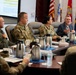 A formal military briefing or conference is taking place in what appears to be a command conference room. Several uniformed military personnel in OCP (Operational Camouflage Pattern) uniforms are seated at a large wooden conference table alongside at least one civilian. A presentation is displayed on a screen in the background, and multiple national and military branch flags are visible.