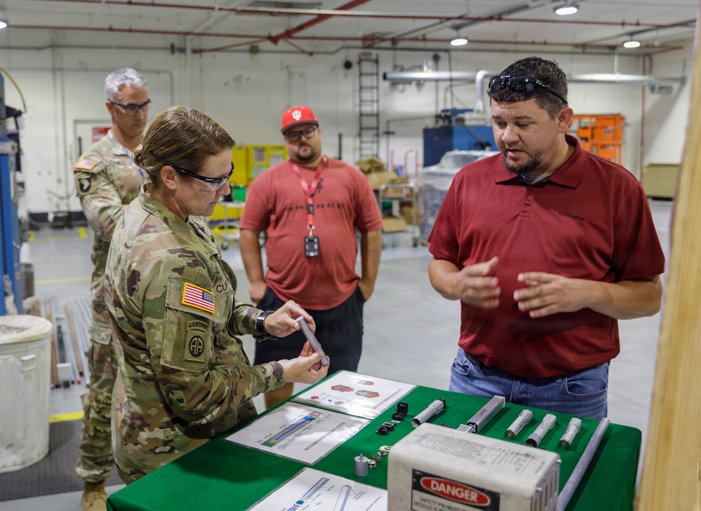 A U.S. Army soldier wearing an Airborne patch and OCP camouflage uniform examines a component at a demonstration table in what appears to be a military maintenance or industrial facility. Two civilian employees in red shirts and one additional soldier observe the demonstration, which features various cylindrical components and technical documents laid out on a green-covered table. A box labeled 'DANGER' is visible on the table, suggesting the items may be safety-critical military equipment.