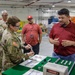 A U.S. Army soldier wearing an Airborne patch and OCP camouflage uniform examines a component at a demonstration table in what appears to be a military maintenance or industrial facility. Two civilian employees in red shirts and one additional soldier observe the demonstration, which features various cylindrical components and technical documents laid out on a green-covered table. A box labeled 'DANGER' is visible on the table, suggesting the items may be safety-critical military equipment.