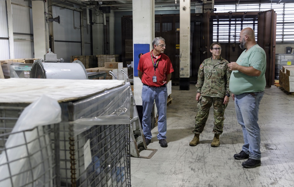 Three individuals are standing in an industrial warehouse or storage facility engaged in conversation. One person is wearing U.S. Army camouflage (OCP) uniform, while the other two are in civilian clothing, one wearing a red shirt with a visible ID badge. The warehouse contains various stored equipment, palletized goods, wire mesh cages, and large cylindrical objects.