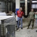 Three individuals are standing in an industrial warehouse or storage facility engaged in conversation. One person is wearing U.S. Army camouflage (OCP) uniform, while the other two are in civilian clothing, one wearing a red shirt with a visible ID badge. The warehouse contains various stored equipment, palletized goods, wire mesh cages, and large cylindrical objects.