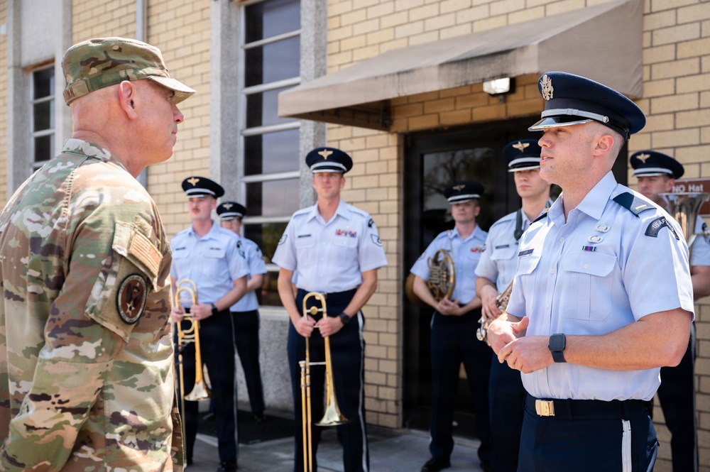 A senior military officer in Army combat uniform (OCP/multicam) is engaged in conversation with a U.S. Air Force officer in service dress uniform. Behind them, a group of Air Force band members stand at ease holding brass instruments including a trombone, trumpet, and French horn. The setting appears to be outside a military building, likely during an official visit or ceremony.