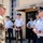 A senior military officer in Army combat uniform (OCP/multicam) is engaged in conversation with a U.S. Air Force officer in service dress uniform. Behind them, a group of Air Force band members stand at ease holding brass instruments including a trombone, trumpet, and French horn. The setting appears to be outside a military building, likely during an official visit or ceremony.