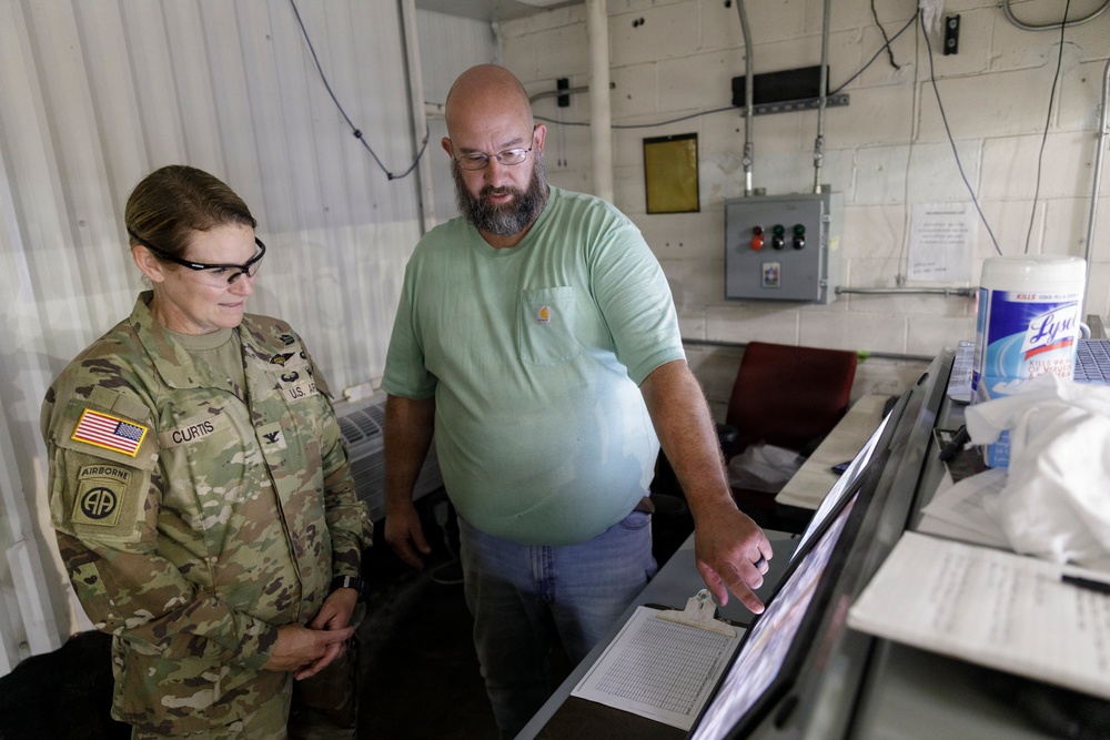 A U.S. Army soldier wearing camouflage uniform with an Airborne patch and the name tag 'Curtis' stands alongside a civilian man in a green t-shirt. They are both looking at a monitor or display screen in what appears to be a control room or operations facility. The civilian is pointing at the screen, suggesting he is explaining or demonstrating something to the soldier.