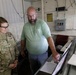 A U.S. Army soldier wearing camouflage uniform with an Airborne patch and the name tag 'Curtis' stands alongside a civilian man in a green t-shirt. They are both looking at a monitor or display screen in what appears to be a control room or operations facility. The civilian is pointing at the screen, suggesting he is explaining or demonstrating something to the soldier.