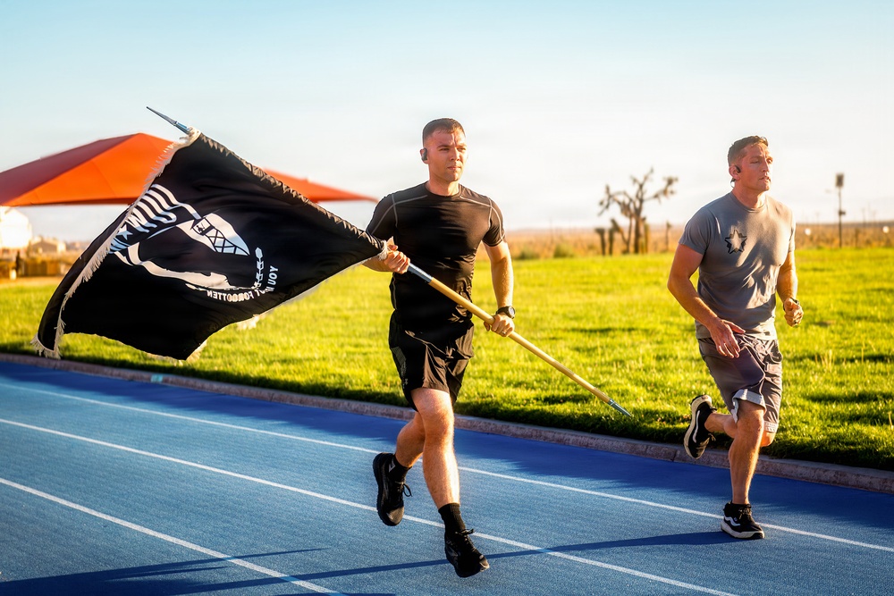 Two men in athletic wear are running on a blue track, with one carrying a black POW/MIA flag on a wooden pole. The flag is billowing in the wind, displaying the recognizable POW/MIA silhouette and the text 'YOU ARE NOT FORGOTTEN' (visible in reverse due to wind direction). The setting appears to be an outdoor athletic facility during golden hour lighting conditions.